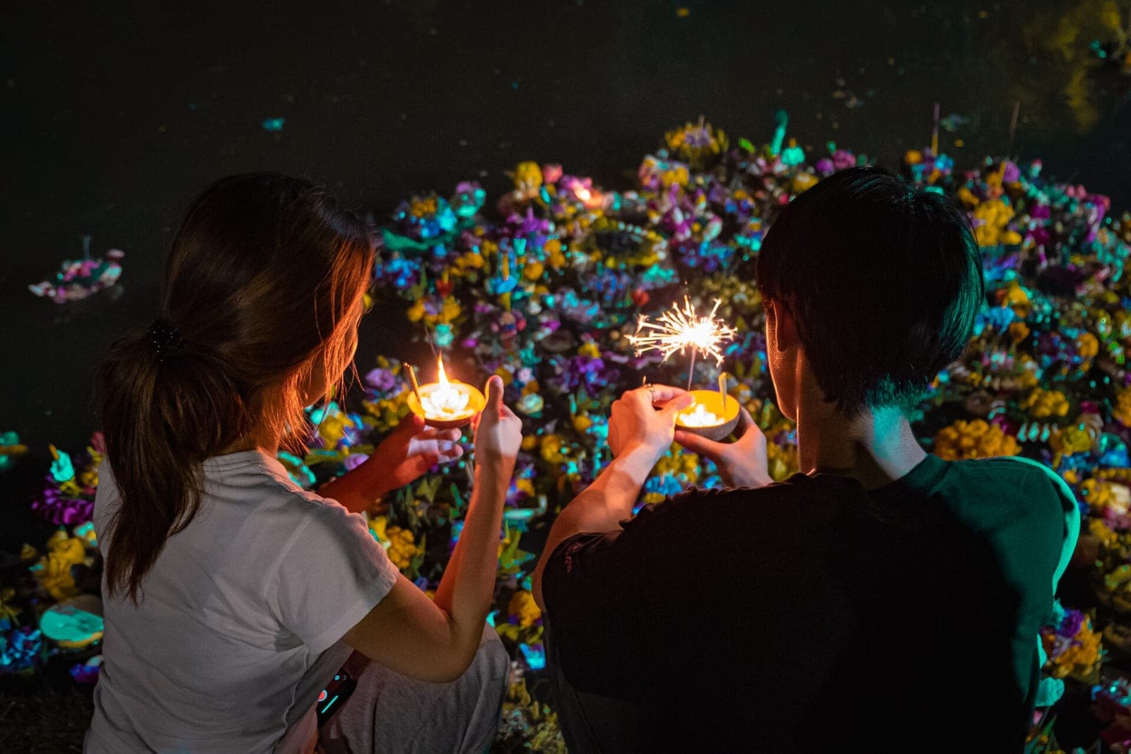 Two people holding candle-lit krathongs beside a pond full of floats during Loy Krathong in Bangkok